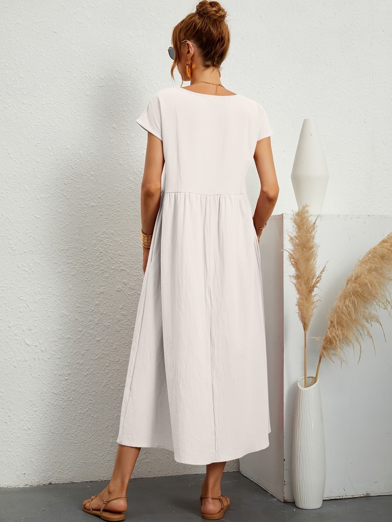 Donna di spalle con abito lungo bianco, sandali beige, accanto a vaso bianco con pampas. Stile estivo, moda casual, abbigliamento femminile.