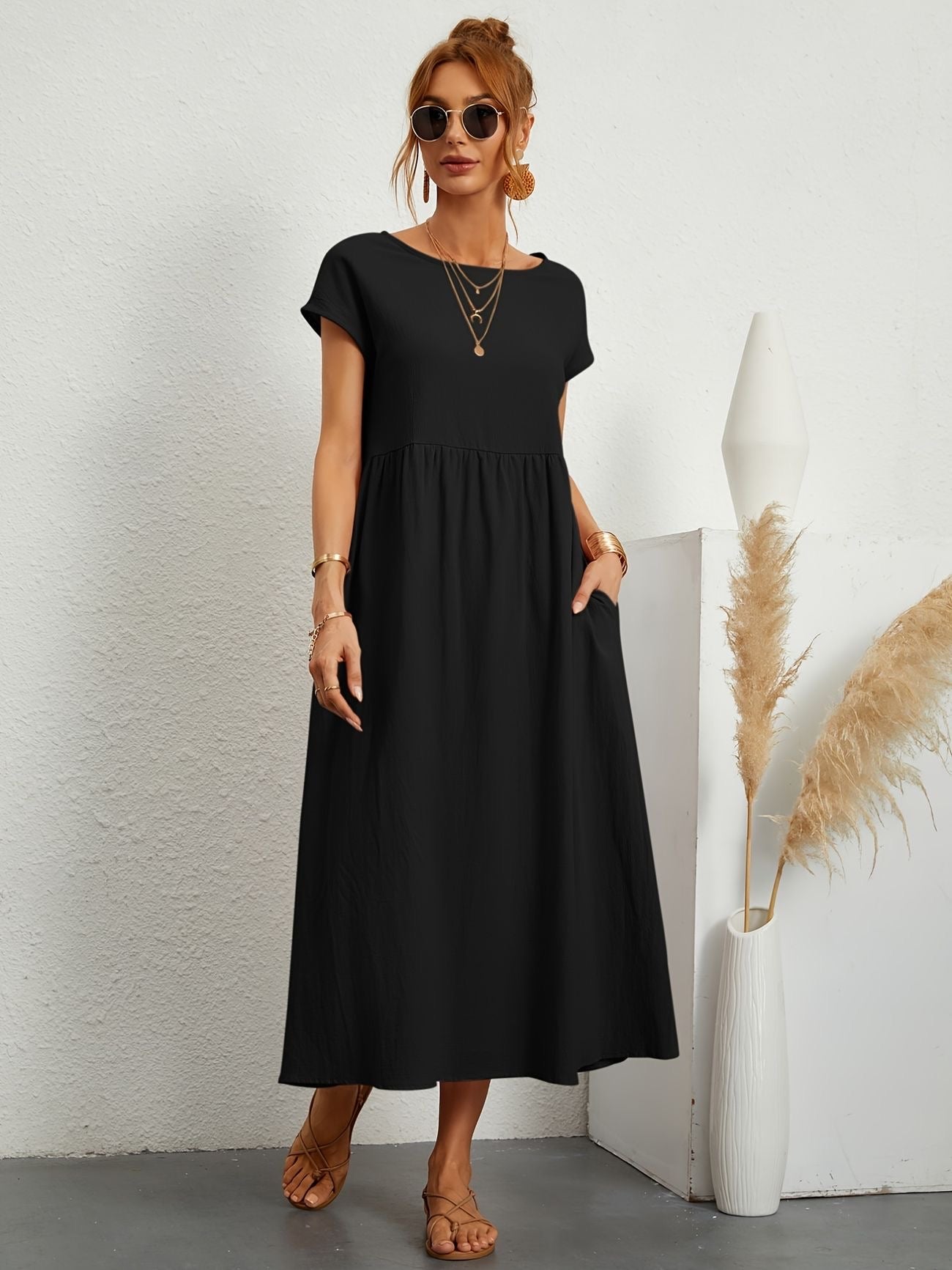 Donna con abito lungo nero, occhiali da sole e sandali, accanto a vaso bianco con pampas. Moda estiva elegante, stile casual chic.