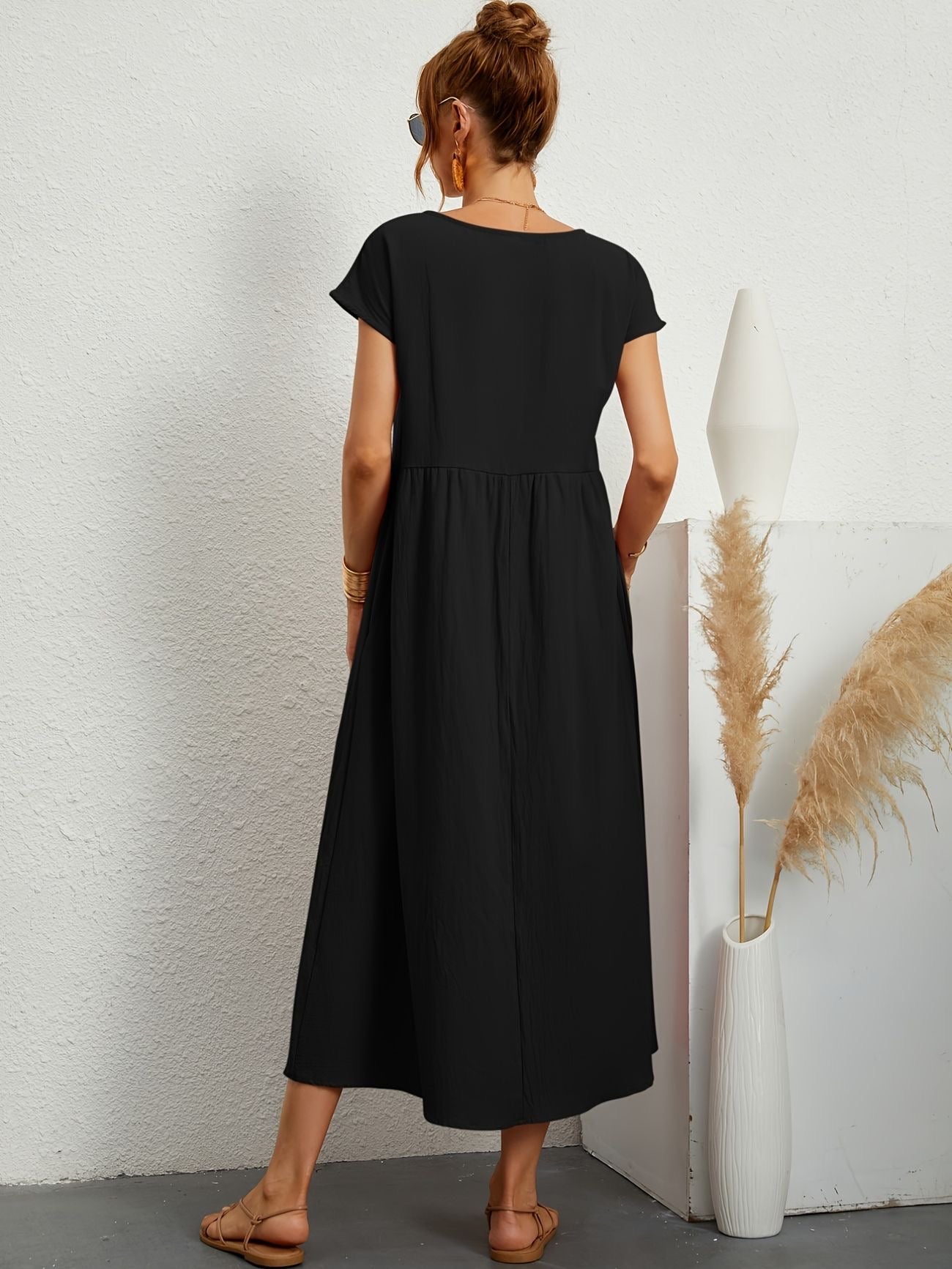 Donna di spalle con abito lungo nero, sandali beige, accanto a vaso bianco con pampas. Moda estiva, abbigliamento casual elegante.