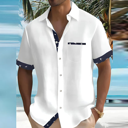Camicia estiva da uomo bianca con dettagli blu, maniche corte, indossata su spiaggia tropicale. Moda mare, abbigliamento casual elegante.