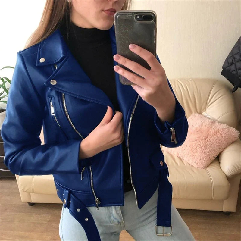 Giacca in pelle blu da donna, stile motociclista, con zip e cintura, indossata sopra un dolcevita nero. Sfondo con divano beige.