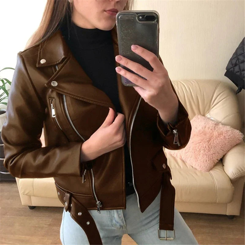 Giacca in pelle marrone da donna, stile motociclista, con zip e cintura, indossata su maglione nero. Look casual su sfondo di salotto moderno.
