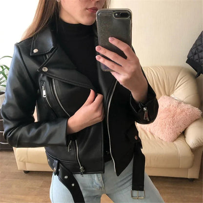 Donna con giacca di pelle nera, maglione nero e jeans chiari, scatta un selfie con smartphone in salotto moderno. Moda autunnale casual.