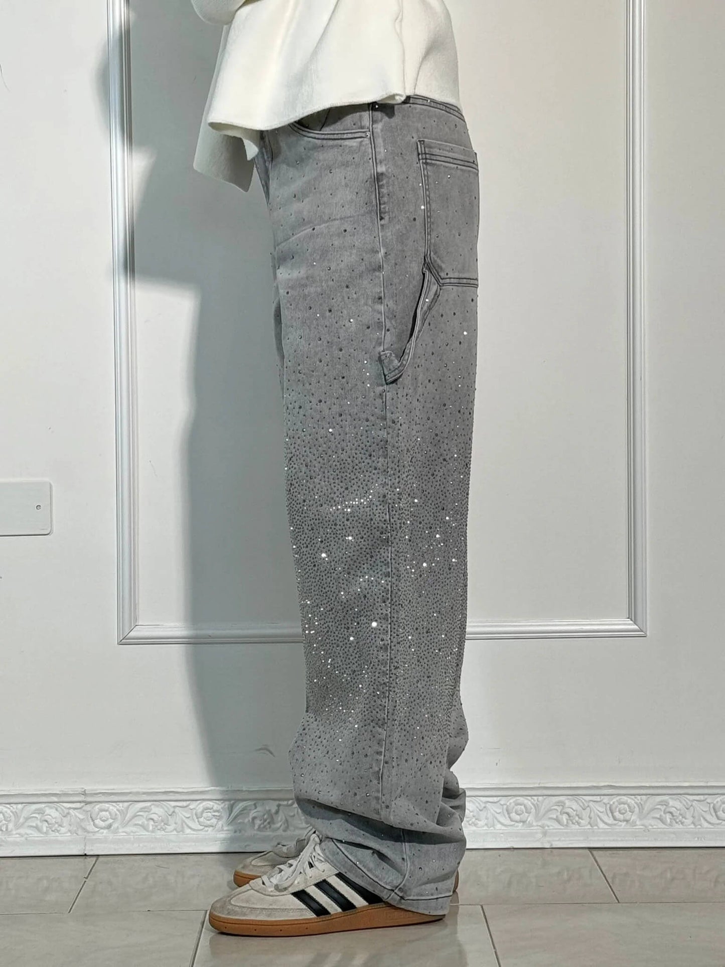 Pantaloni grigi con strass, tasche laterali, indossati con scarpe Adidas bianche e marroni, su sfondo di parete bianca.