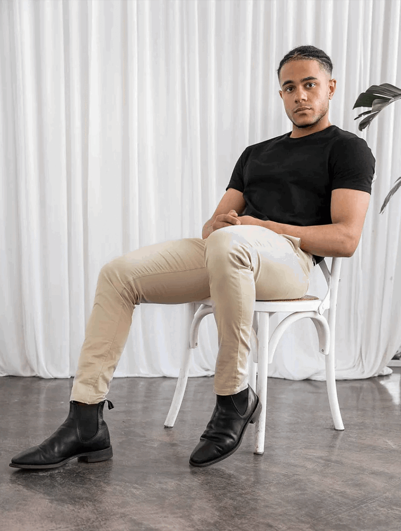 Giovane uomo seduto su sedia bianca, indossa maglietta nera e pantaloni beige, stivali neri eleganti. Sfondo con tenda bianca e pianta decorativa.
