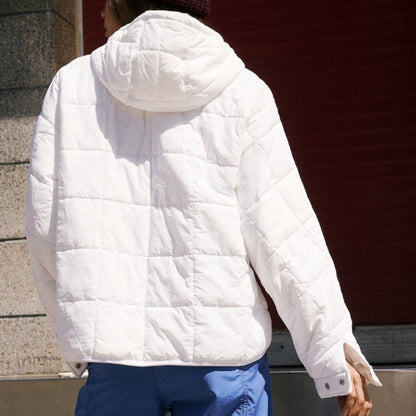 Giacca trapuntata bianca con cappuccio, indossata da una persona di spalle. Sfondo urbano con parete rossa. Moda invernale casual.