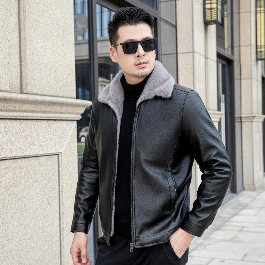 Uomo con giacca di pelle nera e occhiali da sole, posa all'aperto davanti a un edificio moderno. Moda maschile, stile urbano, abbigliamento invernale.