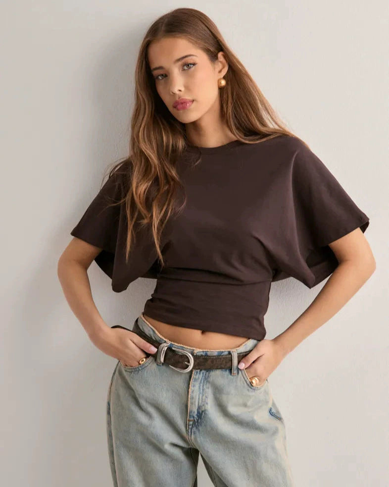Giovane donna con capelli lunghi indossa t-shirt marrone e jeans chiari, posa contro un muro bianco. Moda casual, stile contemporaneo.