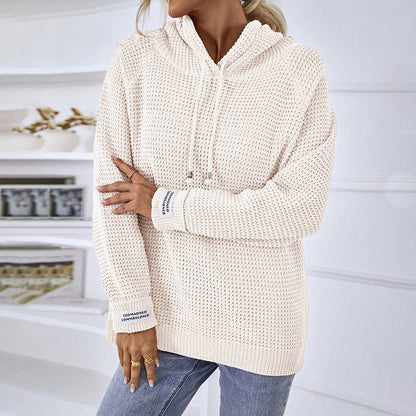 Donna con maglione bianco a maglia grossa, cappuccio e lacci, indossa jeans blu. Sfondo con scaffali e decorazioni moderne. Moda casual autunnale.