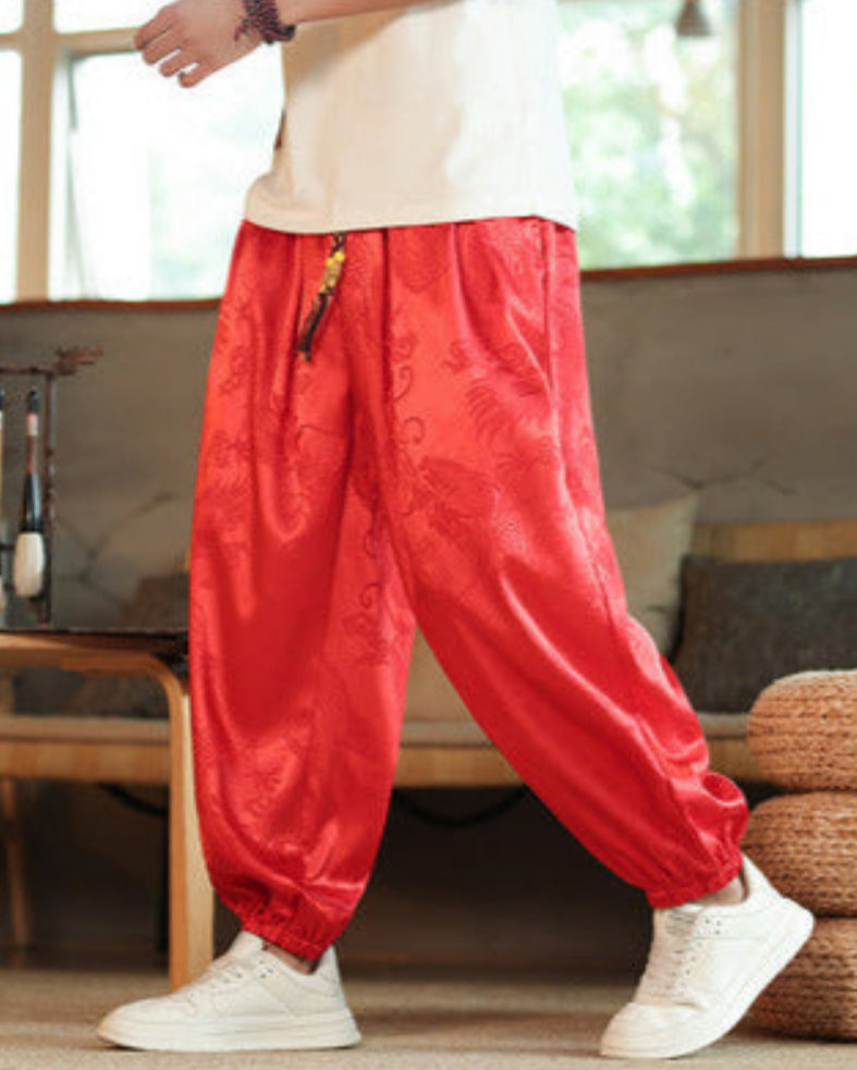 Pantaloni larghi rossi in seta con motivi tradizionali cinesi, indossati da una persona in piedi su un pavimento di legno, interni moderni.