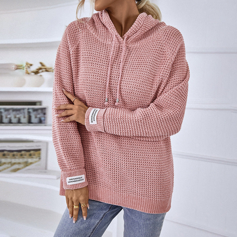 Donna con maglione rosa a maglia grossa, cappuccio e dettagli bianchi, in un ambiente moderno. Abbigliamento casual autunnale, moda femminile.