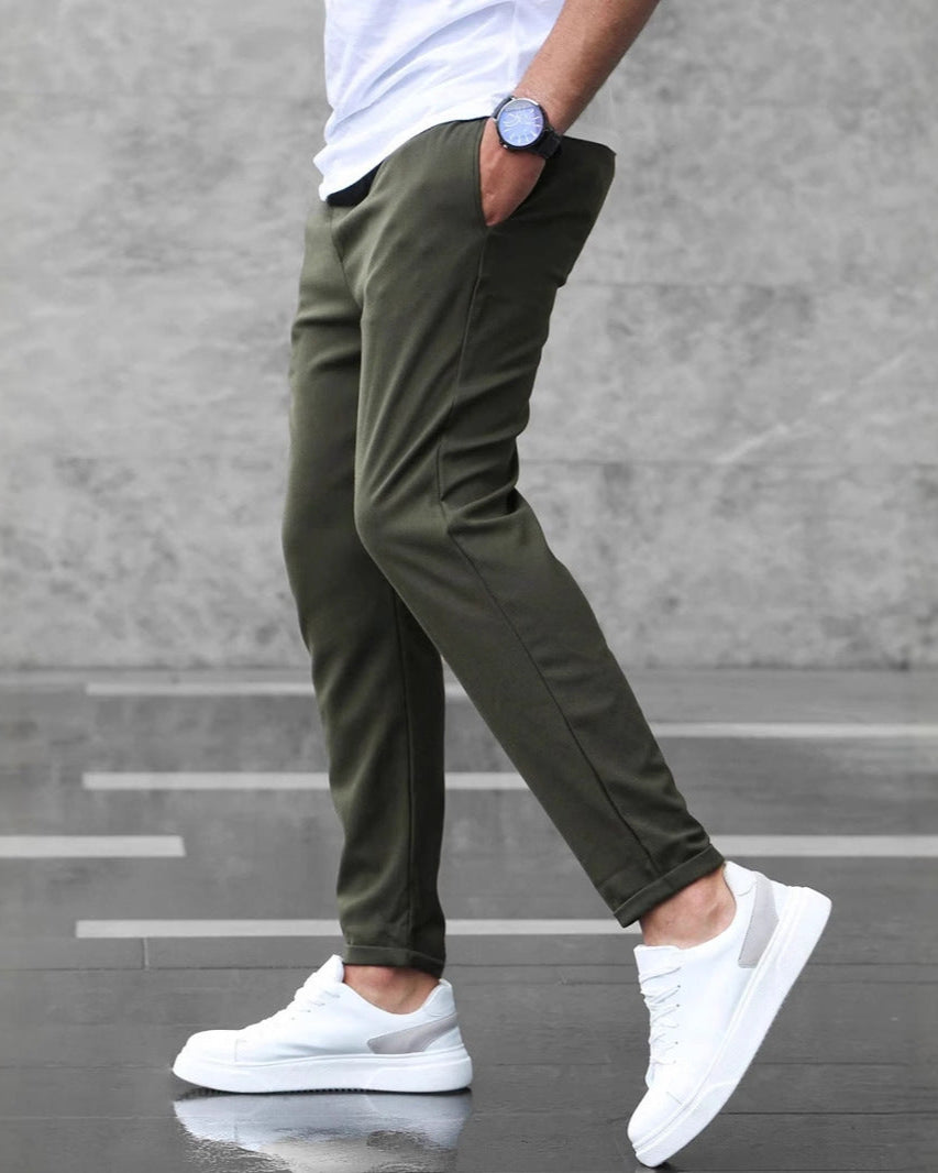 Uomo con pantaloni chino verde oliva, scarpe da ginnastica bianche e orologio blu, cammina su pavimento grigio. Moda casual maschile.