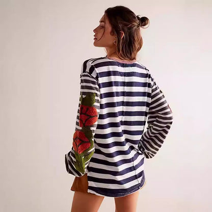 Donna di profilo indossa maglia a righe bianche e blu con maniche lunghe e dettagli floreali colorati. Moda casual, stile moderno.