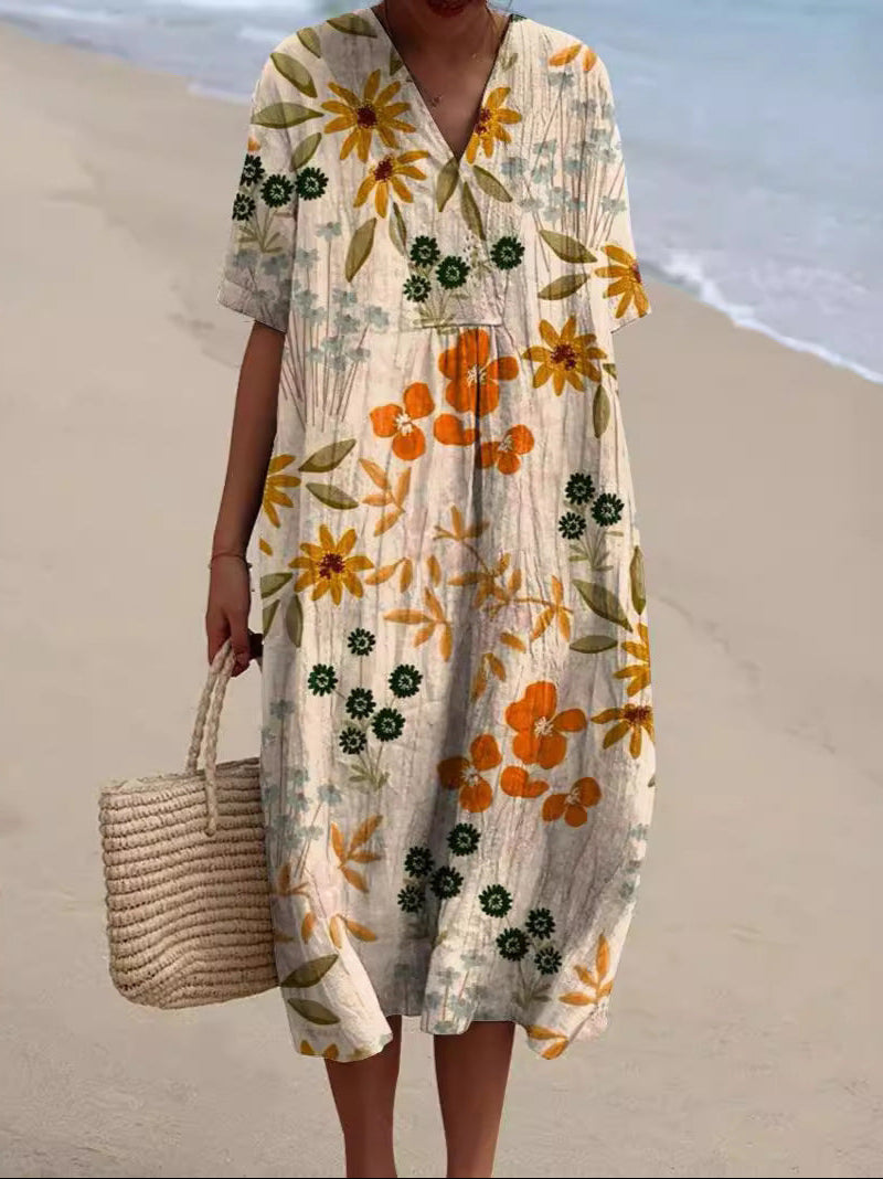Abito estivo floreale lungo con maniche corte, colori arancione e giallo, indossato su spiaggia sabbiosa. Borsa di paglia intrecciata a mano.