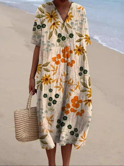 Abito estivo floreale lungo con maniche corte, colori arancione e giallo, indossato su spiaggia sabbiosa. Borsa di paglia intrecciata a mano.