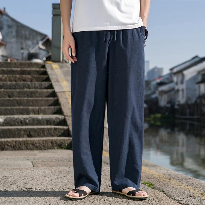 Uomo in pantaloni larghi blu scuro e sandali, in piedi su un marciapiede vicino a un canale, sfondo urbano. Moda casual estiva maschile.