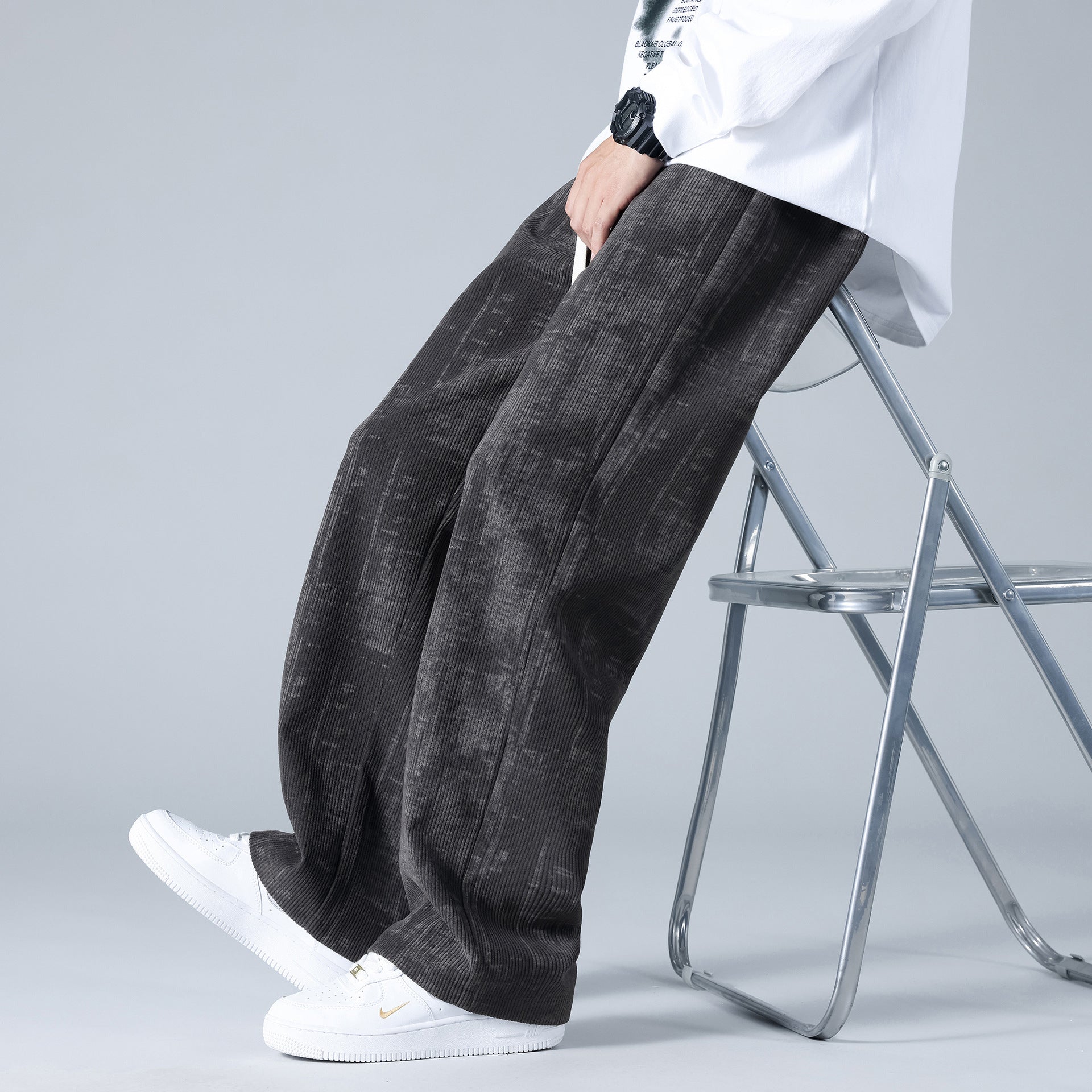 Pantaloni larghi in velluto a coste neri, indossati con scarpe da ginnastica bianche e una camicia bianca, seduto su una sedia pieghevole.
