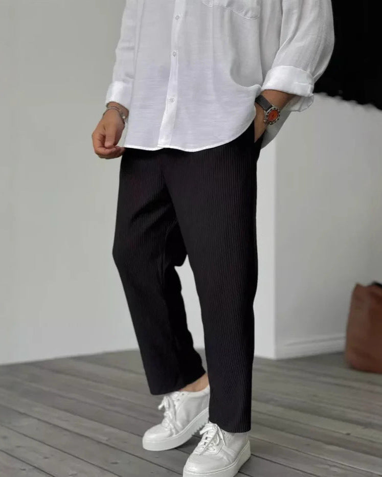 Uomo con camicia bianca e pantaloni neri a pieghe, indossa scarpe da ginnastica bianche su pavimento in legno. Moda casual elegante.