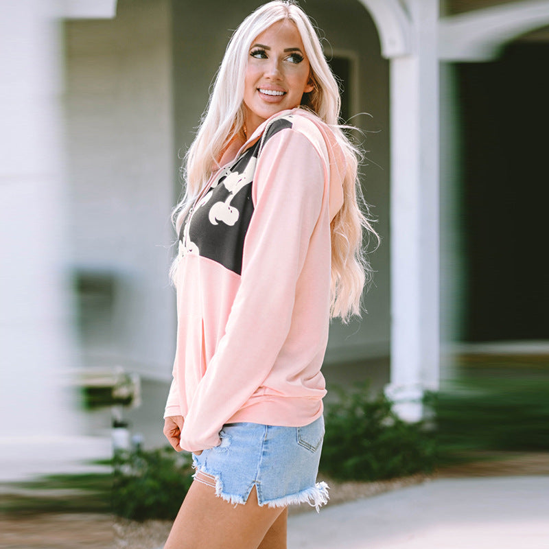 Donna bionda in felpa rosa con cappuccio e shorts di jeans, sorridente all'aperto. Moda casual femminile, abbigliamento estivo trendy.