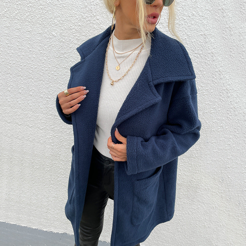 Donna con cappotto blu scuro in pile, maglia bianca e collane dorate, posa contro un muro bianco. Moda autunnale, stile casual chic.