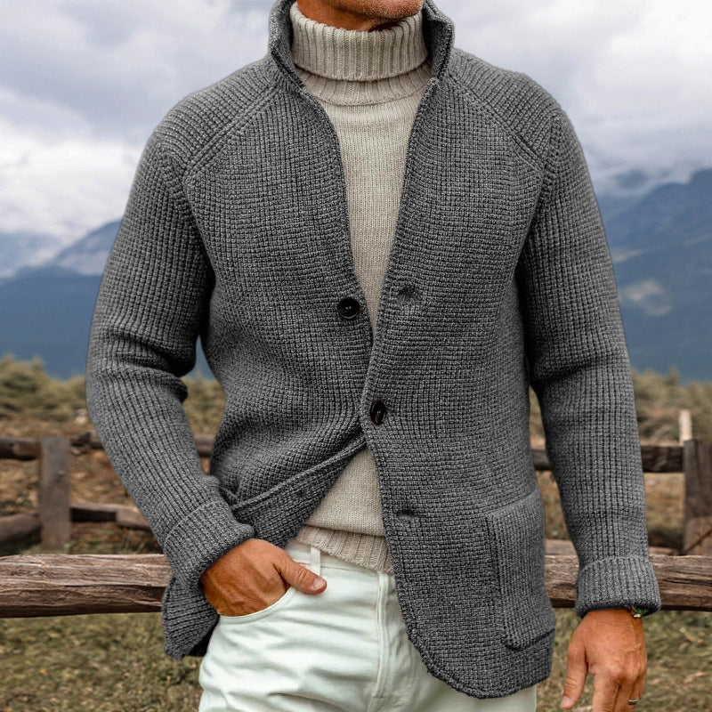 Uomo con maglione grigio a maglia grossa e dolcevita beige, sfondo montano. Moda autunno inverno, abbigliamento casual elegante.