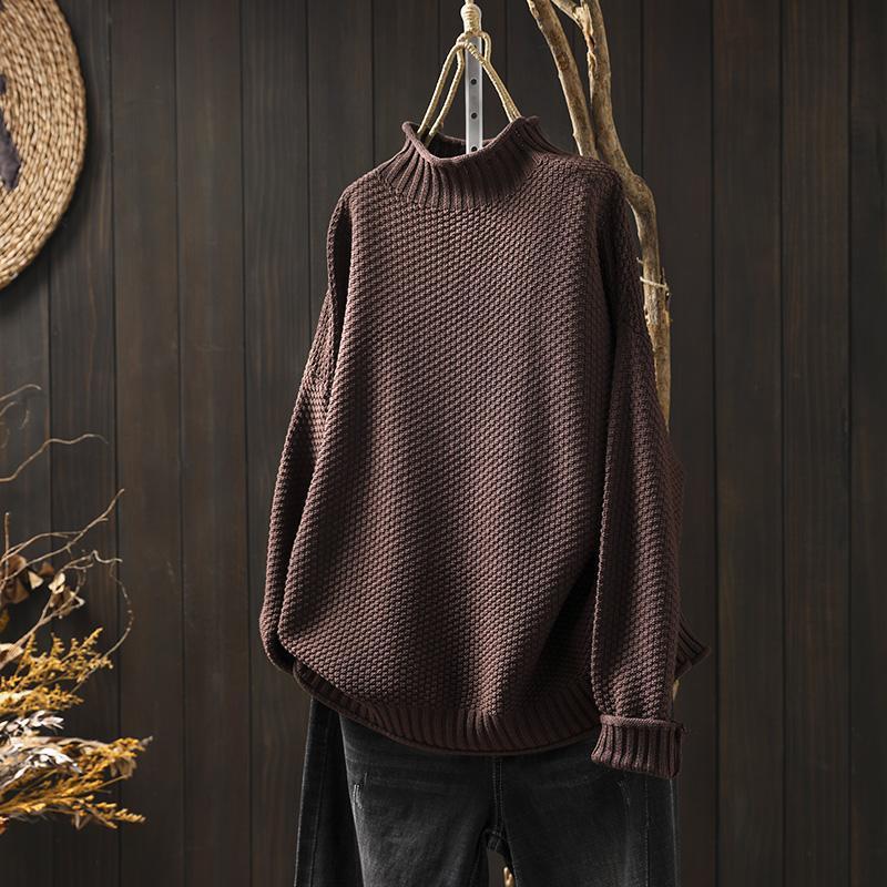 Maglione marrone a collo alto in maglia spessa appeso su sfondo di legno scuro, accanto a rami secchi decorativi. Moda autunnale, abbigliamento caldo.