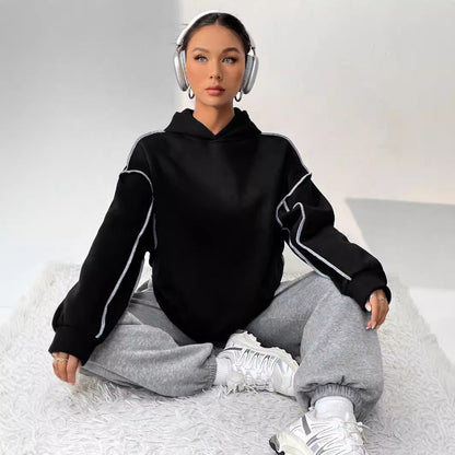 Donna seduta su tappeto bianco, indossa felpa nera, pantaloni grigi, cuffie e sneakers bianche. Moda casual, stile urbano, comfort.