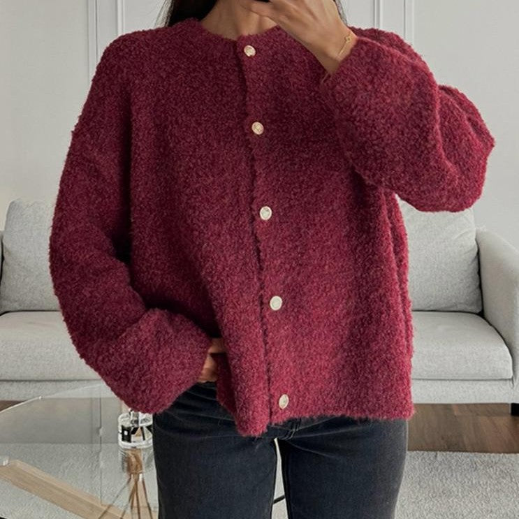 Donna con cardigan rosso borgogna in lana, bottoni bianchi, davanti a un divano grigio. Moda autunno inverno, abbigliamento casual elegante.