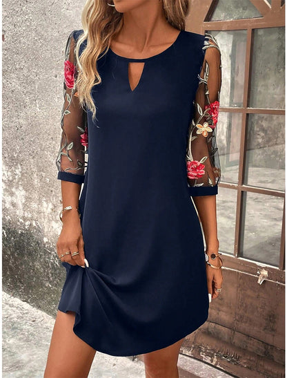 Donna con abito blu scuro, maniche trasparenti ricamate con fiori rossi e bianchi, posa vicino a una finestra. Moda elegante, abbigliamento femminile.