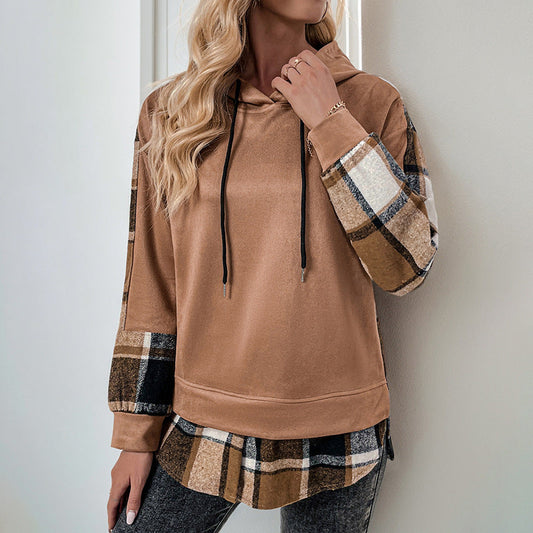 Donna con felpa marrone e maniche a quadri, cappuccio e lacci neri, appoggiata a una parete. Moda casual autunnale, abbigliamento femminile trendy.