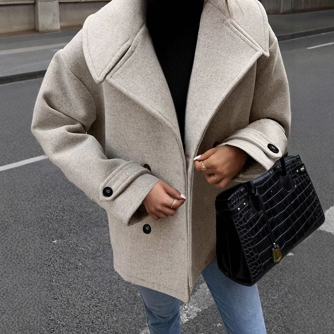 Donna con cappotto beige oversize, maglione nero, jeans, e borsa nera in pelle effetto coccodrillo, cammina su strada cittadina.