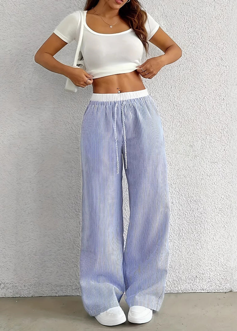 Donna con pantaloni a righe bianche e blu, top bianco corto, posa contro un muro. Moda estiva casual, abbigliamento comodo, stile trendy.