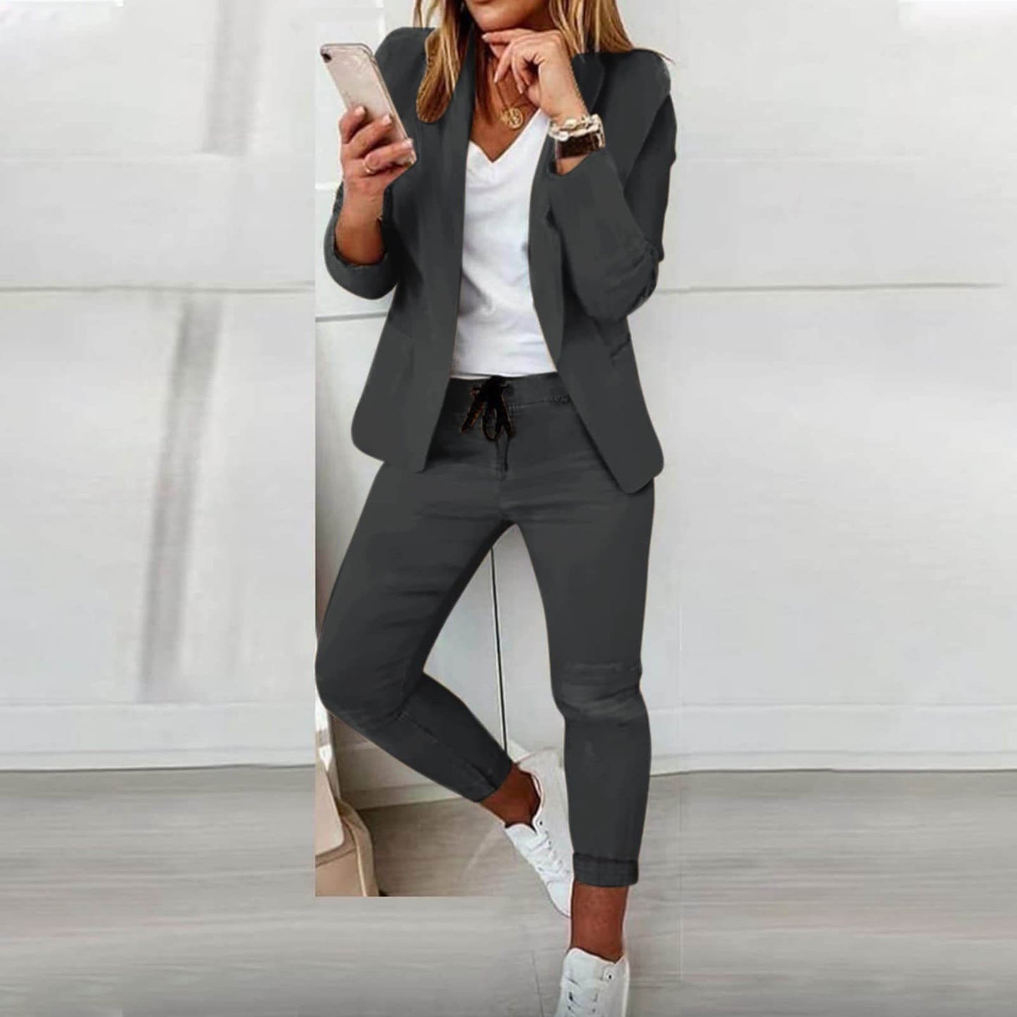 Donna in abbigliamento casual elegante, indossa un completo grigio con giacca e pantaloni, maglietta bianca e scarpe da ginnastica, mentre usa uno smartphone.