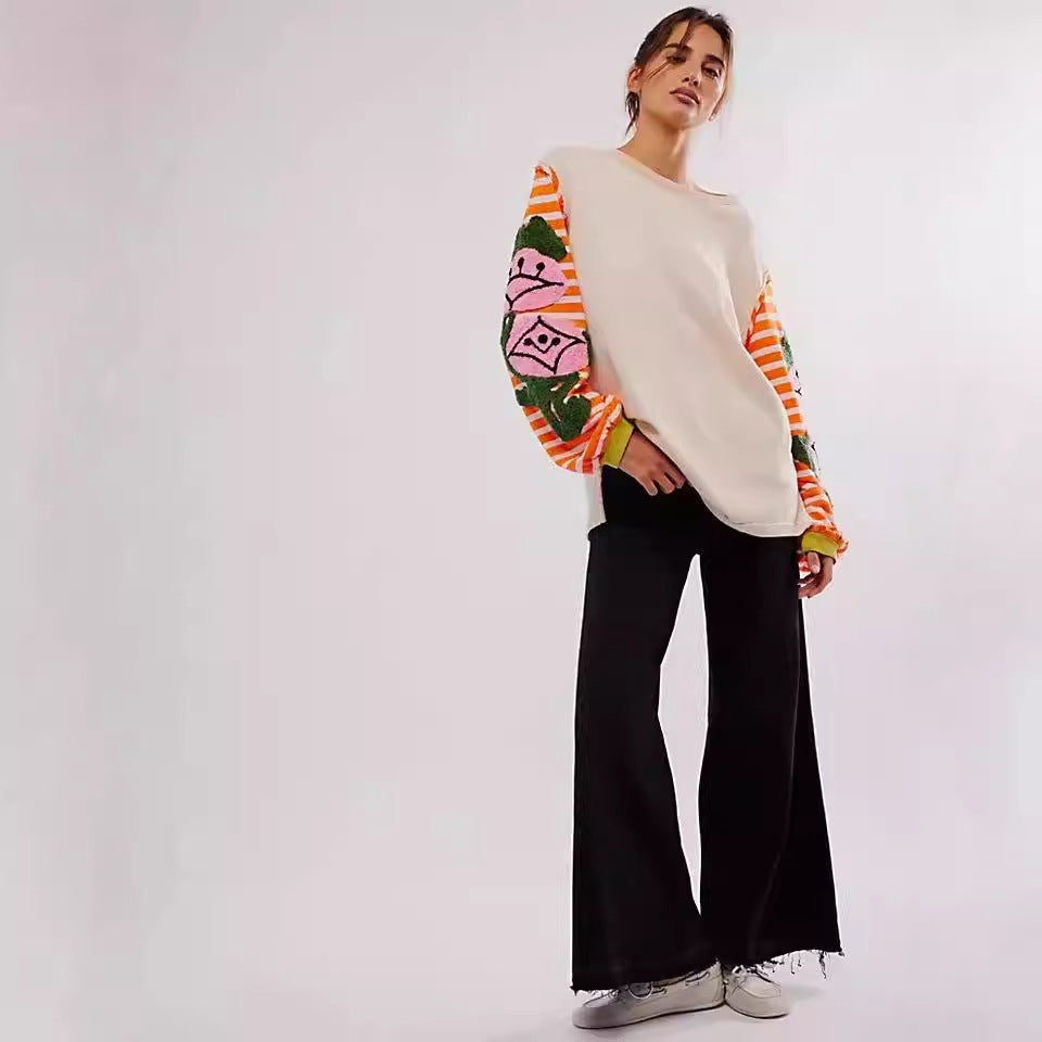 Donna con maglione beige a maniche lunghe colorate e pantaloni neri a zampa, posa su sfondo bianco. Moda autunnale, abbigliamento casual chic.