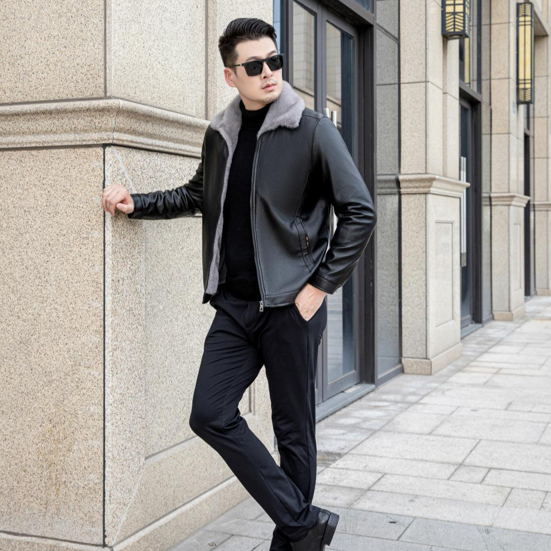Uomo con giacca di pelle nera e occhiali da sole, appoggiato a un edificio moderno. Moda maschile urbana, stile elegante e contemporaneo.