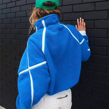 Donna con giacca in pile blu e cappello verde, appoggiata a muro nero. Moda autunno, abbigliamento casual, stile urbano.
