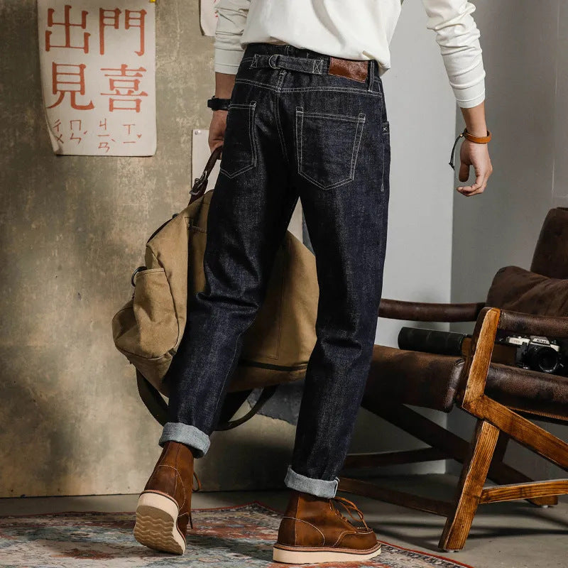 Uomo in jeans scuri e stivali marroni, con maglione bianco, tiene una borsa beige. Sfondo con sedia in legno e poster cinese. Moda casual maschile.
