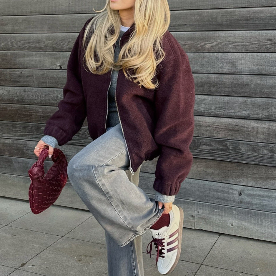 Donna con giacca viola, jeans grigi e scarpe da ginnastica bianche, tiene una borsa intrecciata rossa. Sfondo in legno grigio. Moda casual autunnale.