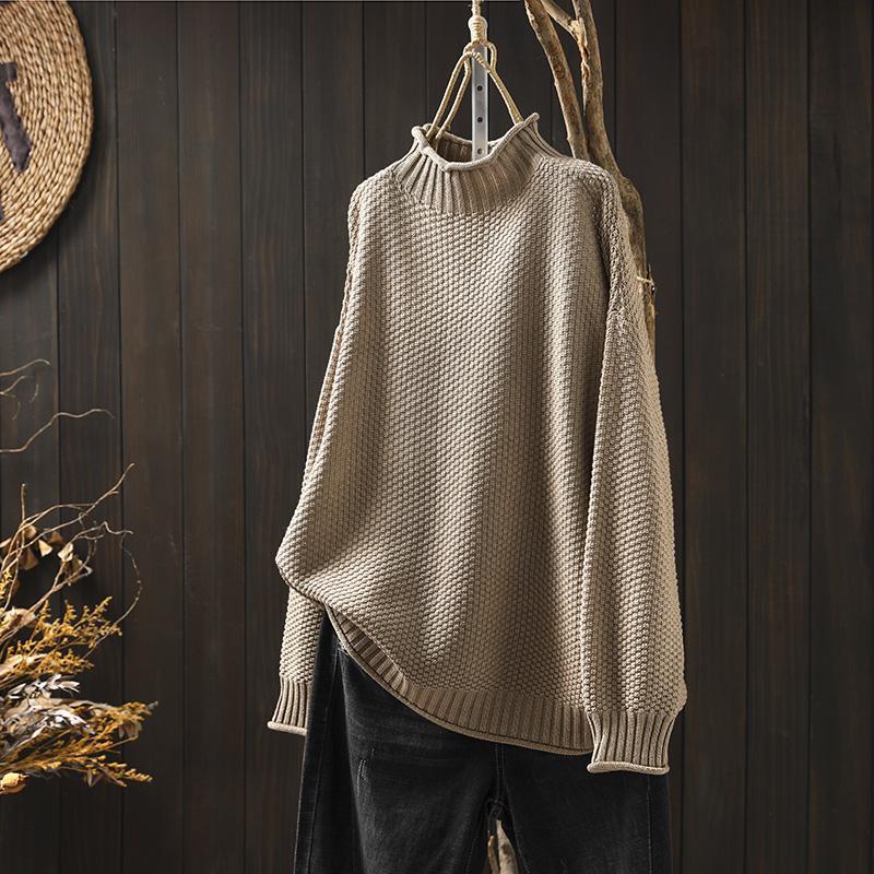 Maglione a collo alto beige in maglia a trama grossa, appeso su sfondo di legno scuro, accanto a decorazioni naturali. Moda autunnale elegante.