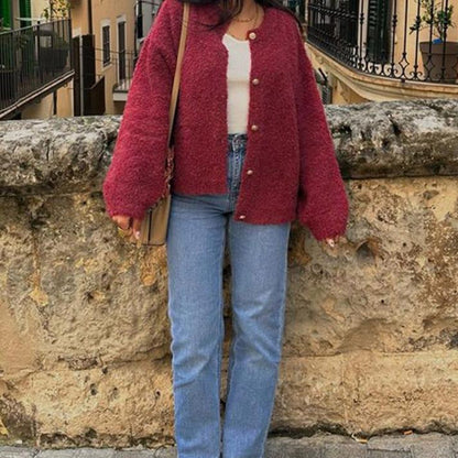 Donna con cardigan rosso e jeans blu davanti a un muro di pietra. Moda autunnale, abbigliamento casual, stile urbano.