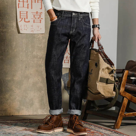 Uomo in jeans scuri e stivali marroni, tiene una borsa beige. Moda casual maschile, abbigliamento autunnale, stile urbano. Sfondo con poster giapponese.