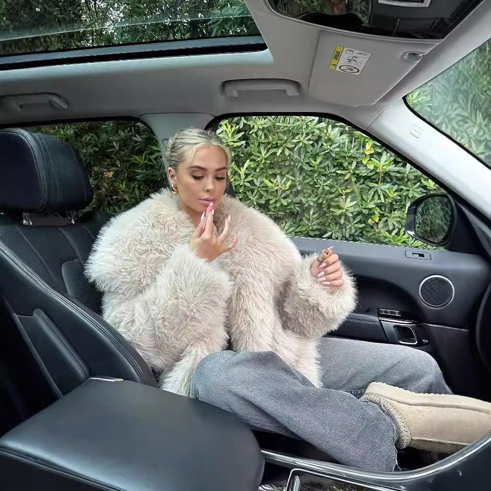 Donna bionda in cappotto di pelliccia beige seduta in auto di lusso, applica rossetto. Interni eleganti, atmosfera sofisticata.