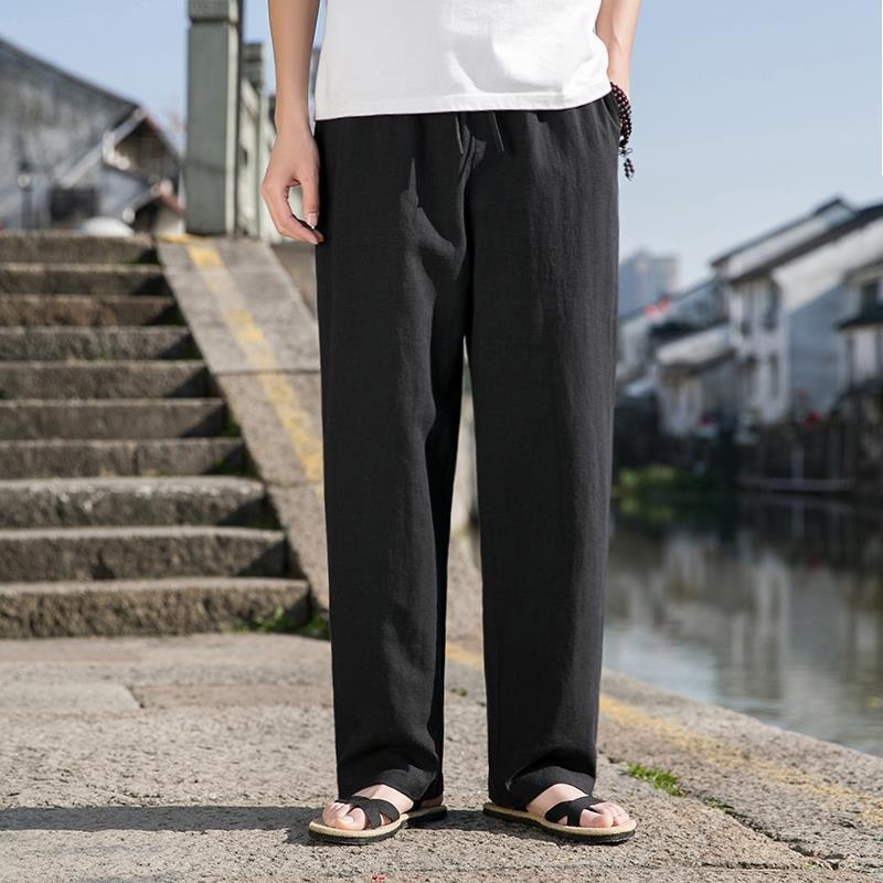 Uomo in pantaloni neri larghi e sandali, vicino a un canale urbano con case tradizionali sullo sfondo. Moda casual estiva maschile.