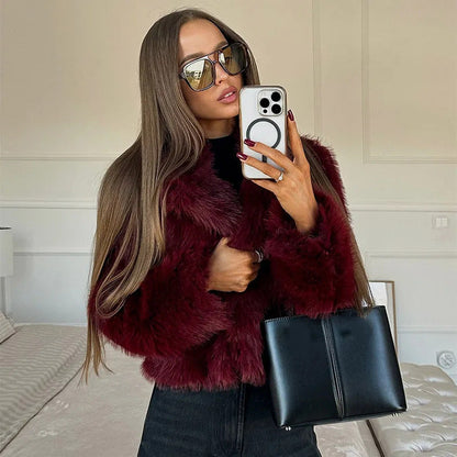 Donna con occhiali da sole, cappotto di pelliccia bordeaux, smartphone e borsa nera in una stanza moderna. Moda autunnale, stile elegante.