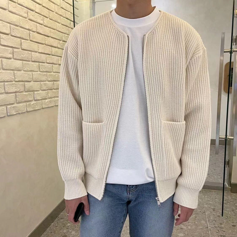 Uomo in cardigan beige a maglia, indossa jeans blu e maglietta bianca, tiene uno smartphone. Sfondo con parete in mattoni bianchi.