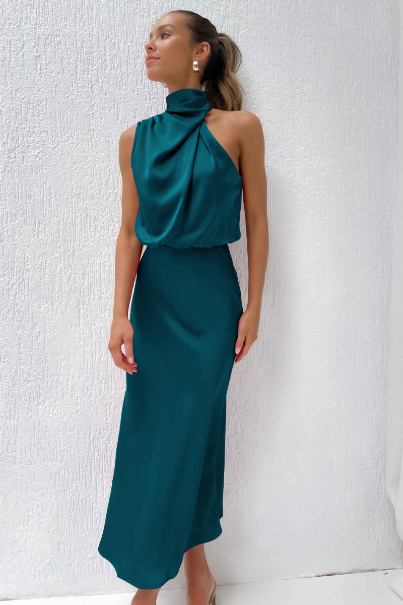 Donna in abito lungo verde smeraldo, elegante e senza maniche, con collo alto drappeggiato, posa contro parete bianca. Moda femminile sofisticata.