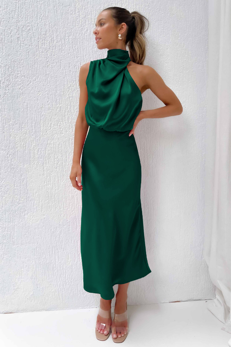 Donna in abito lungo verde smeraldo elegante, senza maniche, con collo alto, posa contro parete bianca. Moda femminile, abbigliamento da sera.