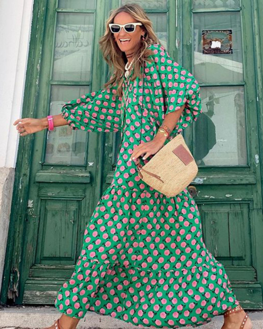 Donna sorridente con abito lungo verde a pois rosa, occhiali da sole bianchi e borsa di paglia, davanti a una porta verde. Moda estiva vivace.