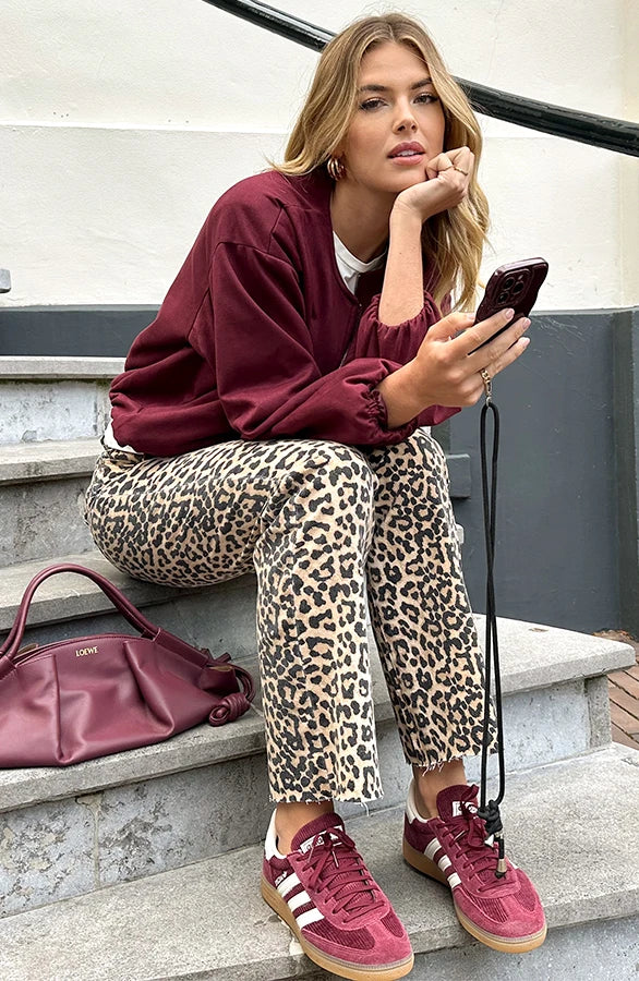 Donna seduta su gradini, indossa maglione bordeaux, pantaloni leopardati, scarpe sportive rosse. Tiene smartphone e borsa abbinata. Moda casual urbana.