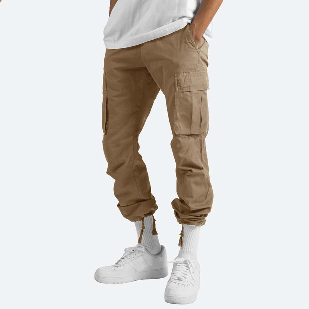 Pantaloni cargo beige da uomo, tasche laterali, abbinati a scarpe da ginnastica bianche e calzini alti. Stile casual e moderno.